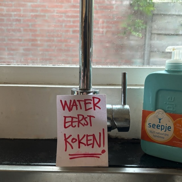 Kraan met een handgetekende notitie eraan die zegt dat water eerst moet koken naast een fles handzeep op een aanrecht met een raam op de achtergrond.
