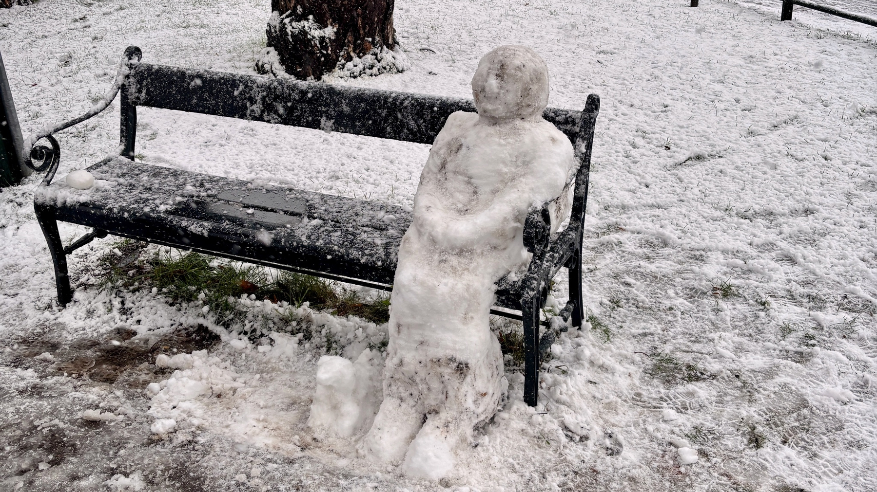 Een sneeuwfiguur zit op een bank en lijkt te ontspannen terwijl sneeuwvlokken neerdalen op de omgeving met gras en een besneeuwde vloer om hem heen.