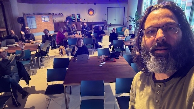 Ik maak een selfie terwijl groep mensen in een ruimte met stoelen en tafels aanwezig zijn en enkele hand omhoog houden in interactie.