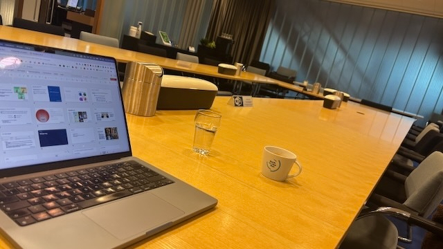 Een laptop staat open op een lange houten vergadertafel naast een glas water en een klein kopje koffie in een goed verlichte vergaderruimte met stoelen rondom.