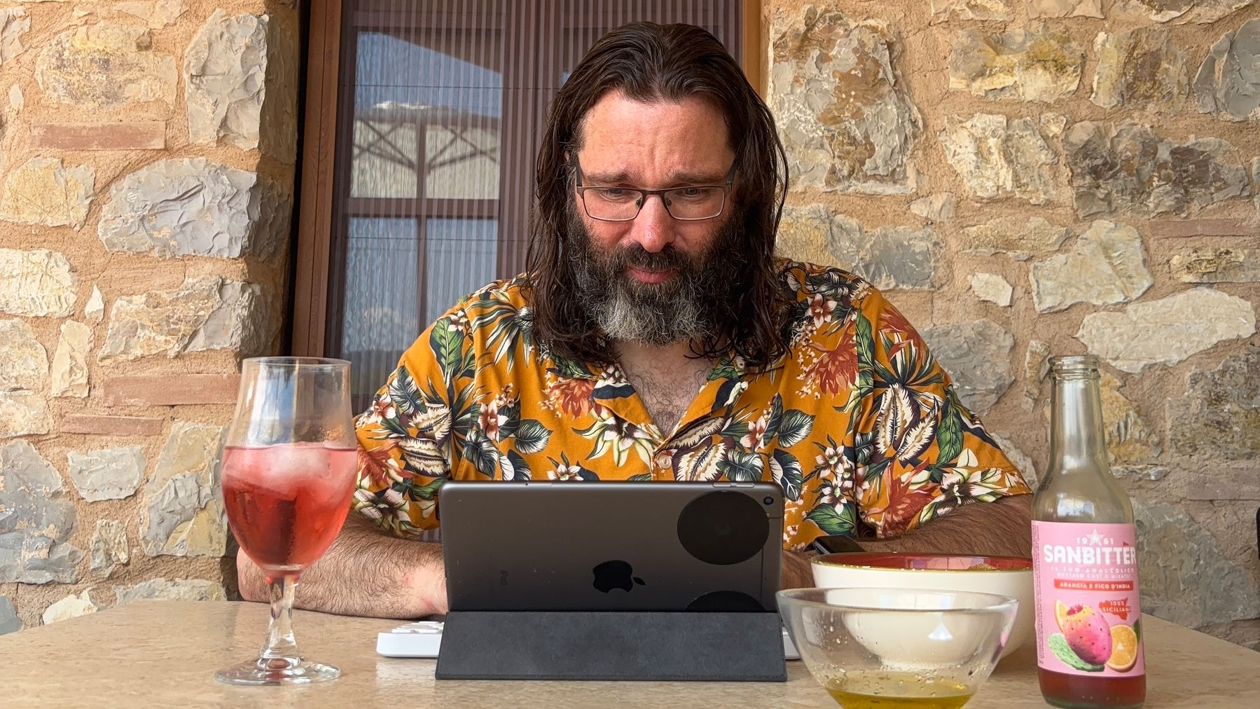Uw auteur kijkt naar een tablet terwijl hij aan een tafel zit met een glas Sanbittèr en wat foccacia en olijfolie.
