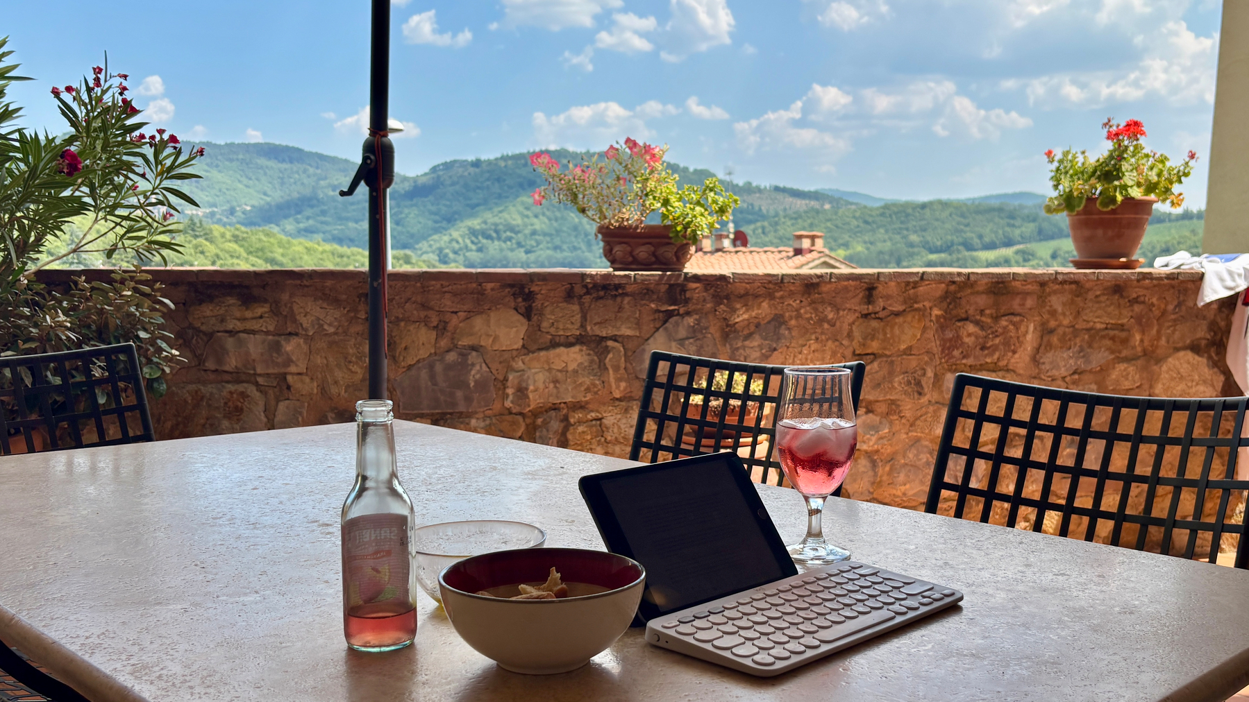 Auto-generated description: Op een terras met uitzicht op groene heuvels staan een tablet met toetsenbord, een glas roséwijn, een flesje en een kom op een tafel.